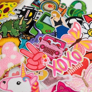 May include: A collection of colourful iron-on patches featuring various designs, including a dinosaur, a butterfly, a jeep, a peace sign, a unicorn, a games console, a pizza slice, and the text "XOXO".