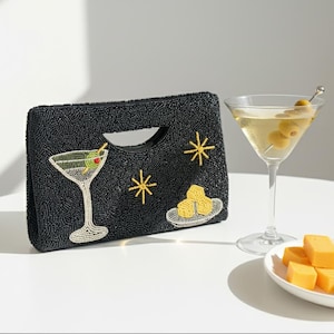 K&ouml;nnte beinhalten: Schwarze, perlenbesetzte Clutch mit Griff, mit einem Martiniglas, Sternen und K&auml;sew&uuml;rfeln. Ein Martiniglas mit Oliven und ein Teller mit K&auml;sew&uuml;rfeln befinden sich daneben. Die Clutch ist etwa 25 cm breit.