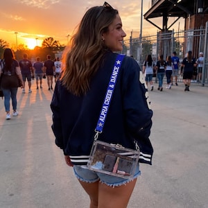 May include: A clear rectangular crossbody bag with a blue strap featuring the white text ALPHA CHEER & DANCE and white star icons. The bag contains a dark brown patterned wallet, a smartphone, and a small tube of lip gloss inside the transparent shell.