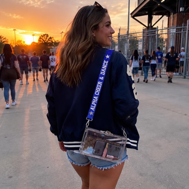 May include: A clear rectangular crossbody bag with a blue strap featuring the white text ALPHA CHEER & DANCE and white star icons. The bag contains a dark brown patterned wallet, a smartphone, and a small tube of lip gloss inside the transparent shell.