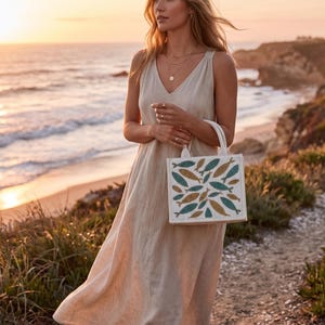 May include: A woman wearing a beige linen dress and holding a white tote bag with a fish pattern in teal and gold. The background shows a beach at sunset, with the ocean and cliffs visible.