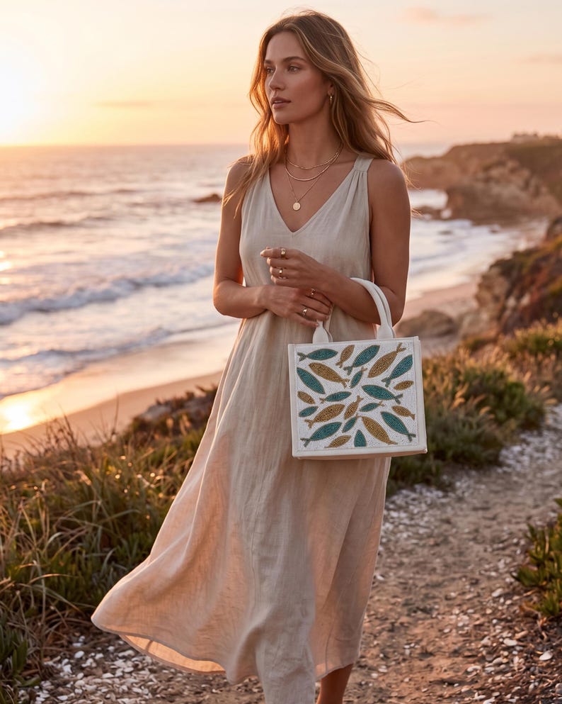 May include: A woman wearing a beige linen dress and holding a white tote bag with a fish pattern in teal and gold. The background shows a beach at sunset, with the ocean and cliffs visible.