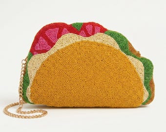 May include: A taco-shaped clutch bag covered in small beads. The design features a golden-yellow shell, green lettuce, red tomatoes, and cream-colored cheese. A thin gold-tone metal chain strap is attached to the side of the beaded handbag.