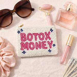 May include: An assortment of beauty and fashion accessories, including a beaded white pouch with the words "BOTOX MONEY" in pink, sunglasses, a pink face roller, perfume, and lip gloss.
