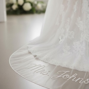 Pode incluir: Um véu de noiva branco com detalhes delicados de renda e o texto bordado "Mrs. Johns" em escrita elegante. O véu é transparente e esvoaçante, com uma borda ondulada. A imagem captura um close-up da parte inferior do véu.