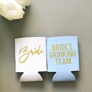 May include: Two white foam beverage insulators. One says "Bride" in gold glitter lettering with a small heart. The other says "Bride's Drinking Team" in gold glitter lettering.