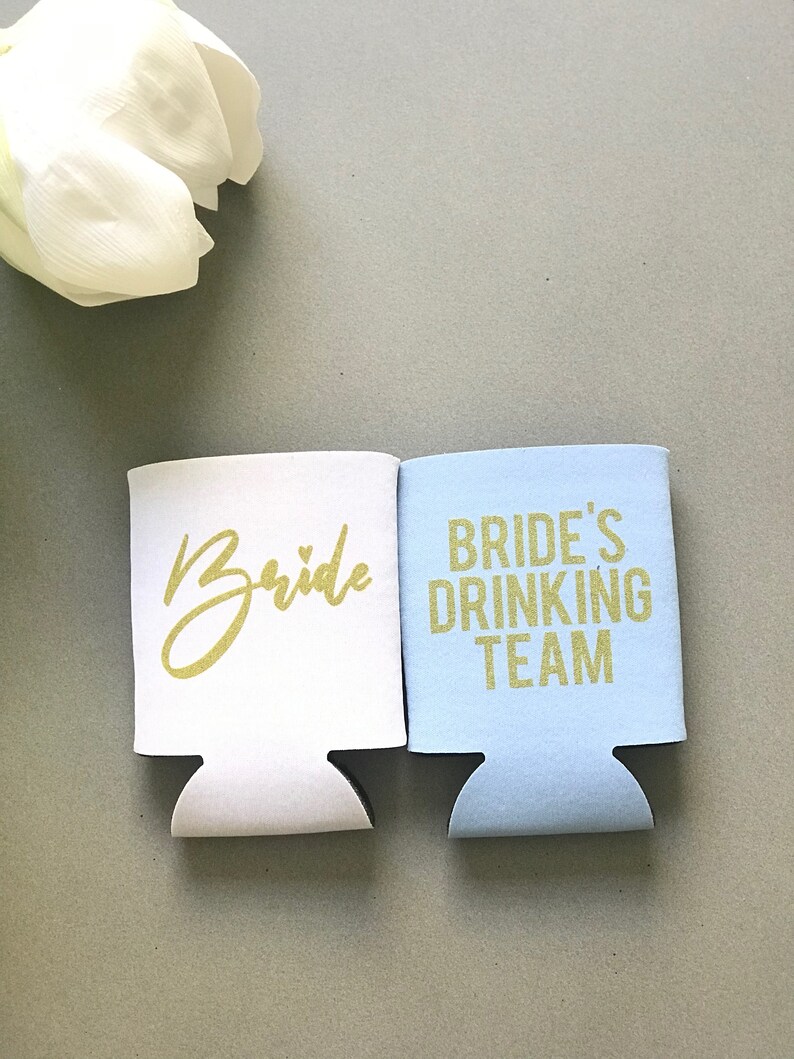 May include: Two white foam beverage insulators. One says "Bride" in gold glitter lettering with a small heart. The other says "Bride's Drinking Team" in gold glitter lettering.