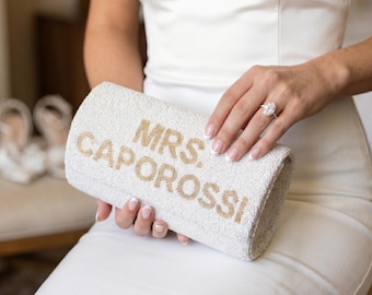 May include: A white beaded clutch purse featuring the text MRS. CAPOROSSI in gold beads. The rectangular bag has a textured surface and is held in place, showcasing the detailed beadwork and the clear lettering on the front panel.