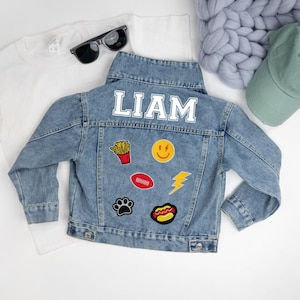 May include: A blue denim jacket with the name "LIAM" embroidered on the back. The jacket has several iron-on patches, including a smiley face, a lightning bolt, a football, a hot dog, french fries, and a paw print.