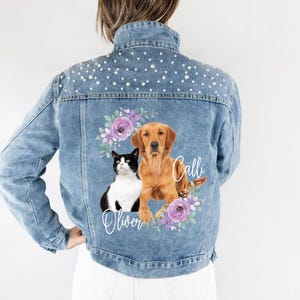 Può includere: Una giacca di jeans blu con decorazioni di perle sul colletto e sulle spalle. Il retro della giacca presenta un design floreale con un gatto e un cane, con i nomi "Oliver" e "Calli" scritti sotto.
