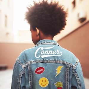 May include: Light blue denim jacket with the name "Conner" in white script across the back. The jacket features patches including a red football, a yellow lightning bolt, a yellow smiley face, and a french fries graphic.