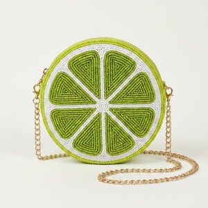 May include: A round, beaded crossbody bag shaped like a lime slice. The design features vibrant lime green segments outlined in white beads, creating a citrus fruit pattern. It includes a detachable gold-tone metal chain strap with lobster clasps.