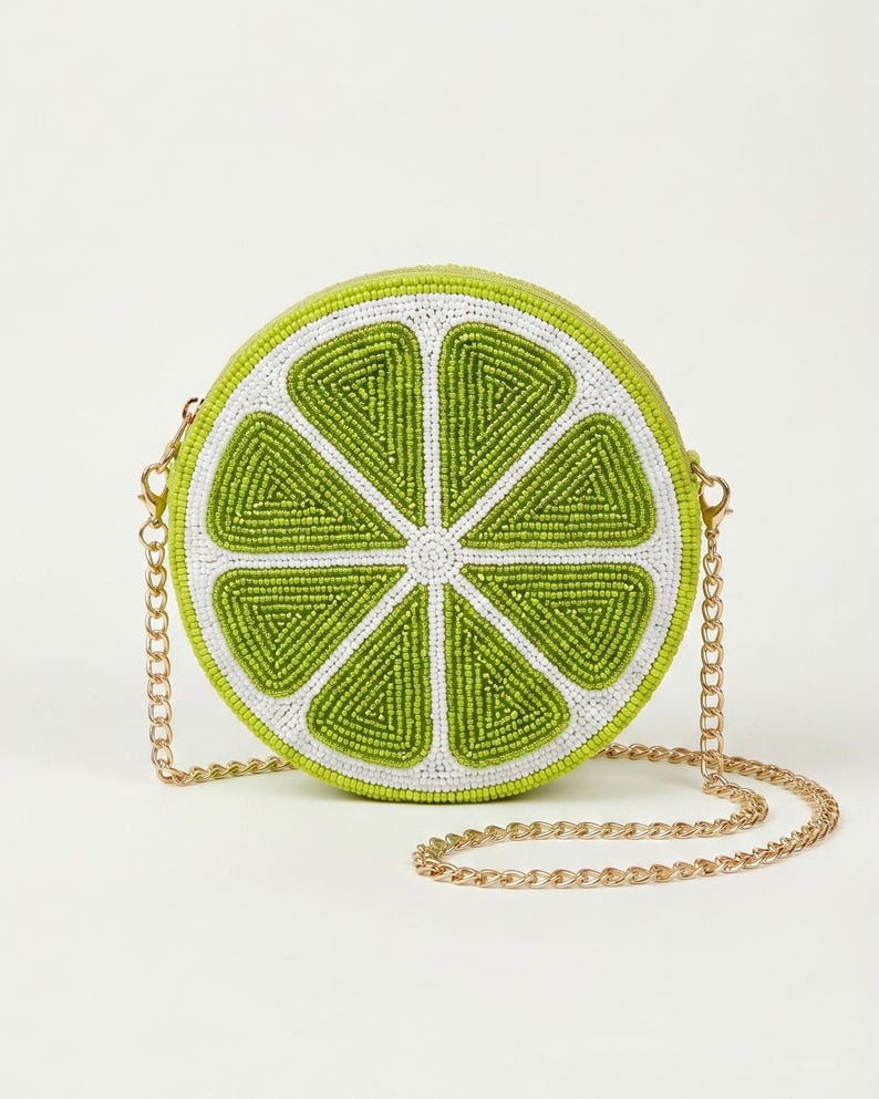 May include: A round, beaded crossbody bag shaped like a lime slice. The design features vibrant lime green segments outlined in white beads, creating a citrus fruit pattern. It includes a detachable gold-tone metal chain strap with lobster clasps.
