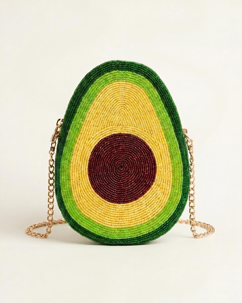 May include: A crossbody bag shaped like a halved avocado, covered in small beads. The design features a dark brown center pit, surrounded by concentric rings of light yellow, bright lime green, and a dark green outer edge, with a gold chain strap.