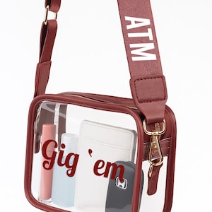 A maroon crossbody bag with a clear front pocket, displaying various items such as a wallet, keys, and a phone.