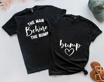 May include: Two black short-sleeved t-shirts. One shirt features the white text THE MAN BEHIND THE BUMP. The other shirt features the white text bump with a heart symbol underneath. The shirts are displayed on a plain surface with floral accents.