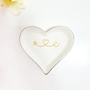 A heart-shaped dish with the word "M" and a heart symbol on it, placed on a white background next to a yellow flower.