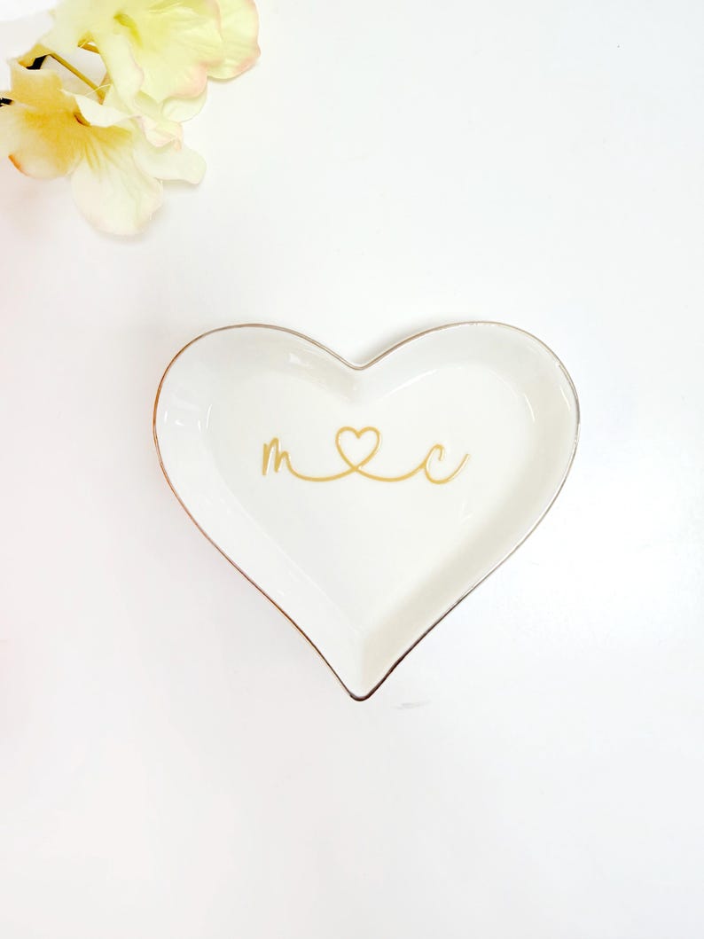 A heart-shaped dish with the word "M" and a heart symbol on it, placed on a white background next to a yellow flower.