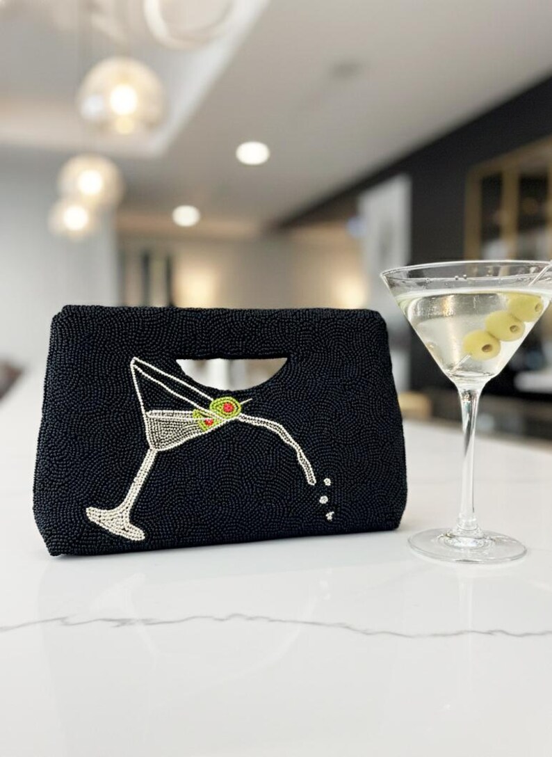 K&ouml;nnte beinhalten: Schwarze, perlenbesetzte Clutch mit Martini-Glas-Design. Die Clutch hat einen Griff und ein Martini-Glas mit Oliven. Ein Martini-Glas mit Oliven befindet sich neben der Clutch.