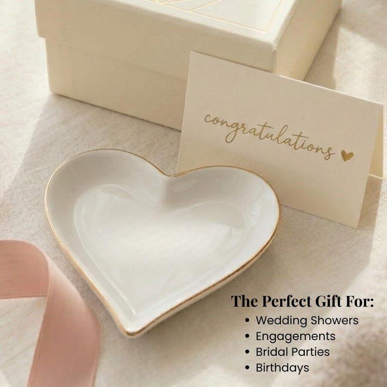 May include: A white heart-shaped ceramic trinket dish with a gold-toned rim. Beside the dish is a cream-colored card with the gold text "congratulations" and a small heart symbol, placed in front of a matching cream-colored gift box.