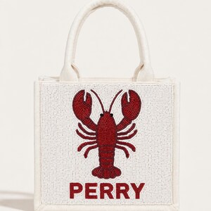May include: A small, off-white tote bag with a beaded design. The design features a red lobster and the name "PERRY" in red letters. The bag has a rectangular shape with a handle.