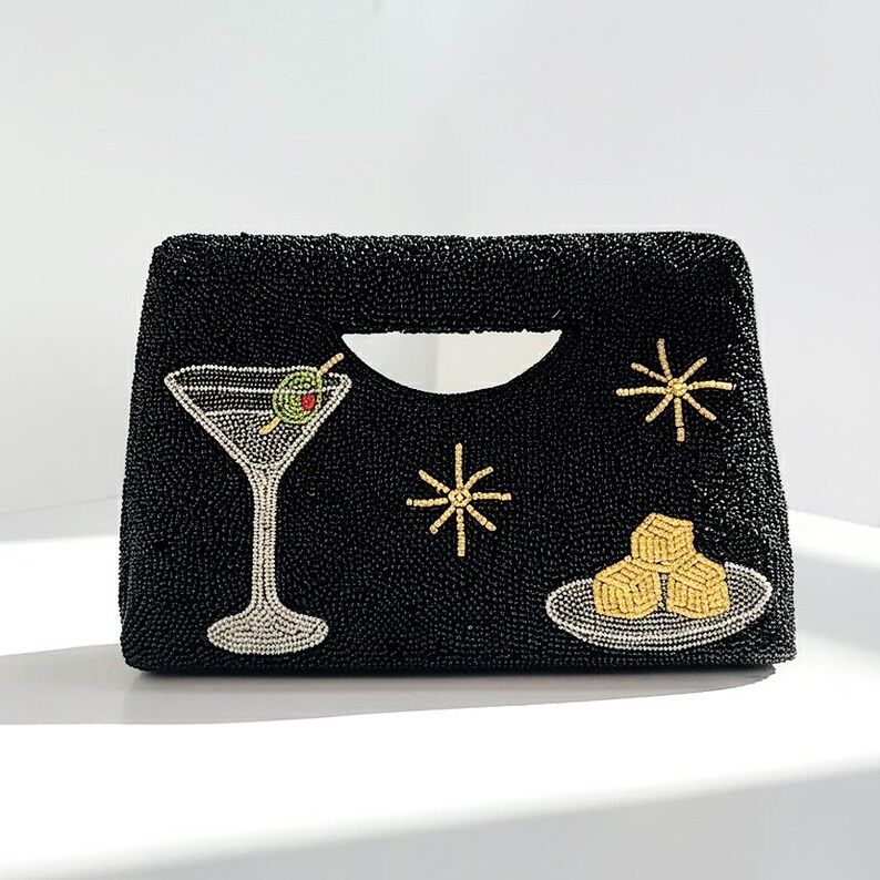 K&ouml;nnte beinhalten: Schwarze, perlenbesetzte Clutch mit Henkel, verziert mit einem Martiniglas, Sternen und einem Teller mit K&auml;sew&uuml;rfeln. Die Clutch hat eine rechteckige Form mit einer geschwungenen Griff&ouml;ffnung.