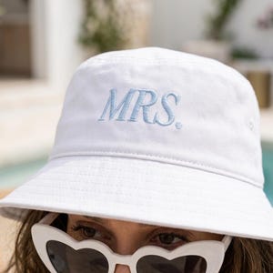 Op de afbeelding: Een witte bucket hat met de geborduurde tekst MRS. in lichtblauw garen. De hoed heeft een gestructureerde kroon en een naar beneden lopende rand. Het ontwerp is eenvoudig en strak, met de nadruk op het contrast tussen de witte stof en de blauwe tekst.