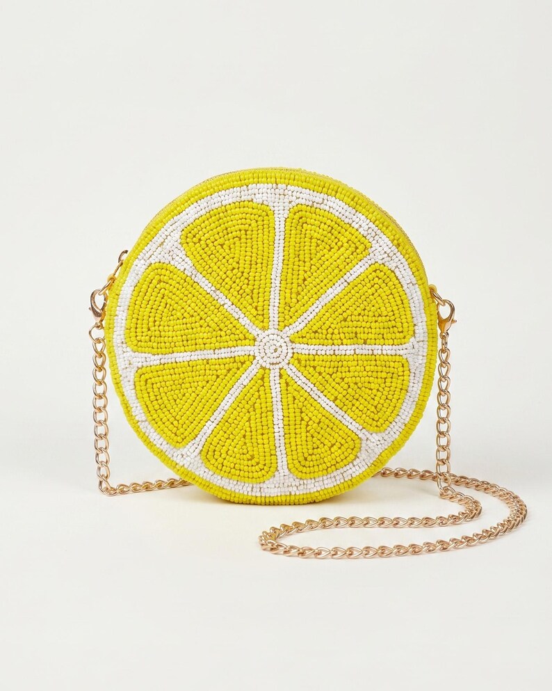 May include: A round crossbody bag shaped like a lemon slice, featuring vibrant yellow and white glass beads arranged in a radial pattern. The bag includes a thin, metallic gold-tone chain strap attached to the sides with small lobster clasps.