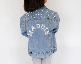 Könnte beinhalten: Eine hellblaue Jeansjacke mit einem Rückenteil, das mit weißen Kunstperlen verziert ist. Der Name MADDIE ist in weißen Großbuchstaben mittig auf den Rücken gestickt, ergänzt durch weitere Perlenakzente an der oberen Passe.