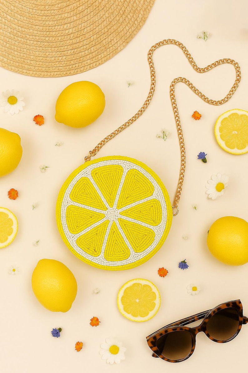 May include: A round, beaded crossbody bag shaped like a lemon slice with yellow and white beads, featuring a gold-tone metal chain strap. The bag is surrounded by whole lemons, lemon slices, small white and orange flowers, and tortoise shell sunglasses.