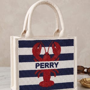 May include: A canvas tote bag with a beaded design. The front features navy and white horizontal stripes with a red lobster and the name "PERRY" in navy. The bag has cream-colored handles and trim.
