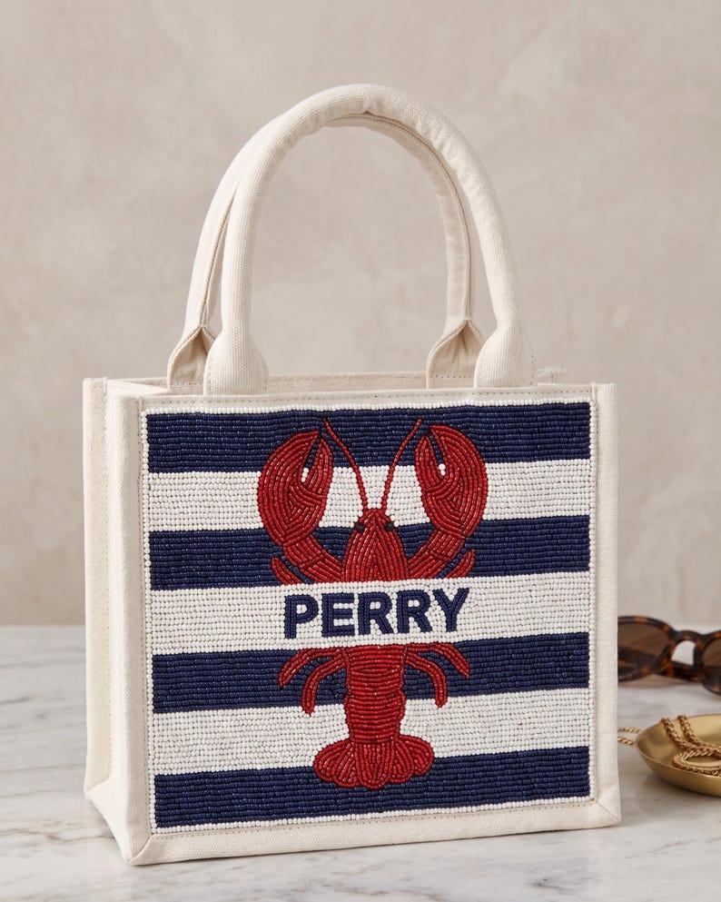 May include: A canvas tote bag with a beaded design. The front features navy and white horizontal stripes with a red lobster and the name "PERRY" in navy. The bag has cream-colored handles and trim.