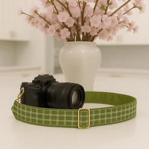 May include: A black digital camera with a lens rests on a green and white plaid camera strap. The strap has a gold-colored buckle. A white vase with pink flowers is in the background.