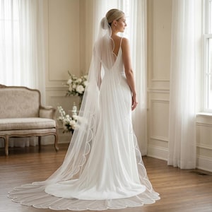 Könnte beinhalten: Eine Braut in einem weißen Hochzeitskleid und Schleier steht in einem hellen Raum. Das Kleid hat ein schlichtes Design mit tiefem Rückenausschnitt. Der lange Schleier hat einen gewellten Rand. Der Raum hat weiße Vorhänge und ein Sofa im Vintage-Stil.