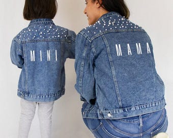Könnte beinhalten: Zwei blaue Jeansjacken mit weißen Perlenverzierungen am oberen Rücken und an den Schultern. Die kleinere Jacke trägt den Schriftzug MINI in Weiß auf dem Rücken, während die größere Jacke den Schriftzug MAMA in Weiß aufgedruckt hat.