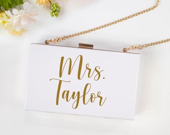 May include: White clutch purse with a gold chain strap. The purse has the text "Mrs. Taylor" written in gold lettering.