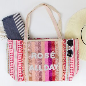May include: A pink and white striped tote bag with the text "ROS&Eacute; ALL DAY" in white glitter. The bag has pink pom-poms on the sides and a blue and white striped beach towel is next to it.