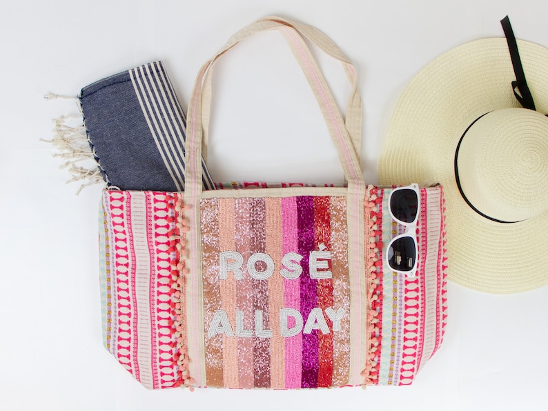 May include: A pink and white striped tote bag with the text "ROS&Eacute; ALL DAY" in white glitter. The bag has pink pom-poms on the sides and a blue and white striped beach towel is next to it.