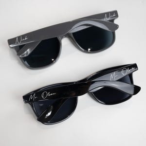 May include: Two pairs of sunglasses with black frames and dark lenses. One pair has the name "Nick" written on the frame, and the other pair has the text "Mr. Olson" written on the frame.