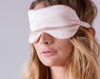 May include: A pale pink satin sleep mask covers the eyes of a person with long, wavy blonde hair.