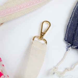 May include: A close-up of a beige canvas strap with a gold metal clasp. The strap is attached to a bag with a pink stripe.