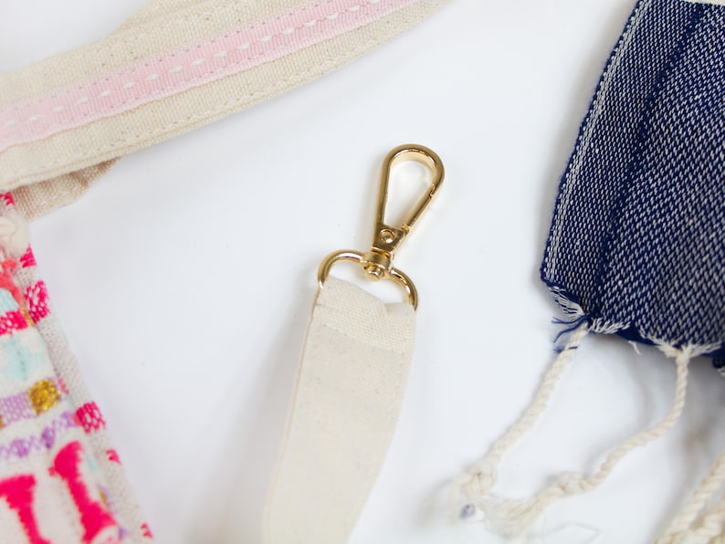 May include: A close-up of a beige canvas strap with a gold metal clasp. The strap is attached to a bag with a pink stripe.
