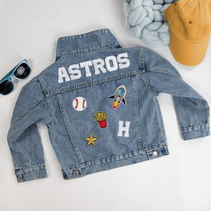 Könnte beinhalten: Eine hellblaue Jeansjacke mit dem weißen Schriftzug ASTROS auf der Rückseite. Die Jacke ist mit gestickten Aufnähern verziert, darunter ein Baseball, eine Rakete, eine Portion Pommes frites, ein goldener Stern und der weiße Buchstabe H.