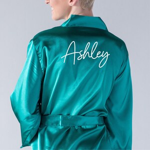 May include: Teal satin robe with a matching belt. The word "Ashley" is written in white script across the back. The robe has long sleeves and a shawl collar.