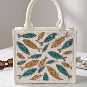 May include: Off-white canvas tote bag with a beaded fish design in teal and gold. The bag has a rectangular shape with a matching off-white handle. The fish pattern is set against a white background.