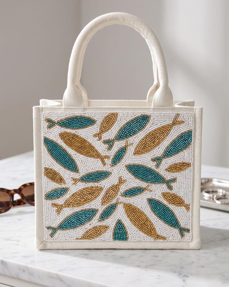May include: Off-white canvas tote bag with a beaded fish design in teal and gold. The bag has a rectangular shape with a matching off-white handle. The fish pattern is set against a white background.