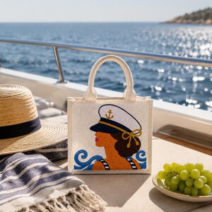 May include: A square-shaped tote bag made of off-white fabric featuring a beaded graphic of a person wearing a white captain's hat with a gold anchor emblem. The design includes blue wave patterns at the bottom and two sturdy top handles.