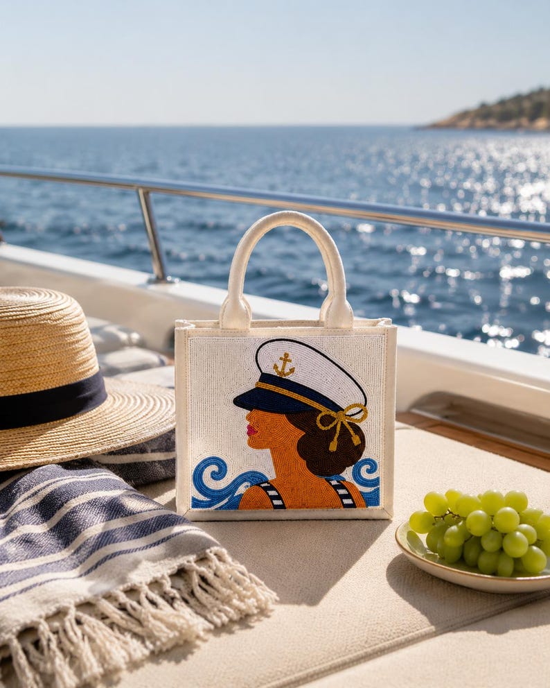 May include: A square-shaped tote bag made of off-white fabric featuring a beaded graphic of a person wearing a white captain's hat with a gold anchor emblem. The design includes blue wave patterns at the bottom and two sturdy top handles.