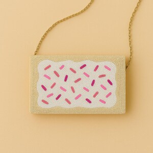 May include: A rectangular beaded clutch bag shaped like a pastry with a tan crust border and a white frosted center. The surface is decorated with small pink and magenta beaded sprinkles and includes a thin gold-toned metal chain shoulder strap.