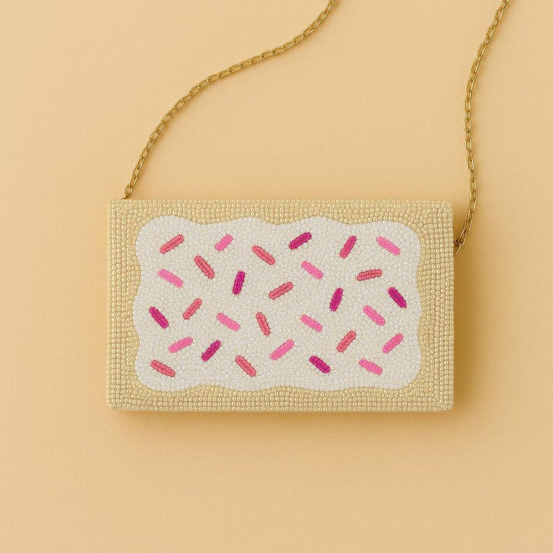 May include: A rectangular beaded clutch bag shaped like a pastry with a tan crust border and a white frosted center. The surface is decorated with small pink and magenta beaded sprinkles and includes a thin gold-toned metal chain shoulder strap.